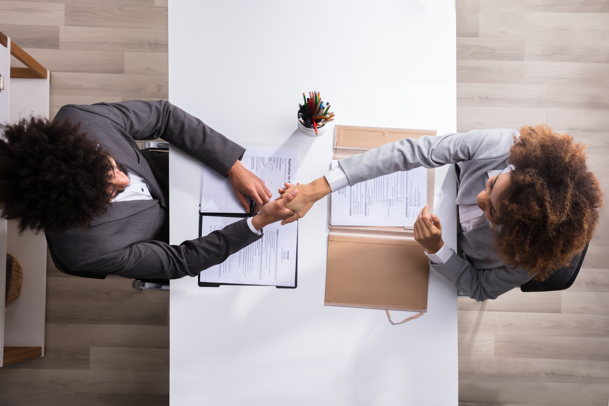 Above angle photo of male manager shaking the hand of a female applicant while sitting at a table. Paperwork on the table.
