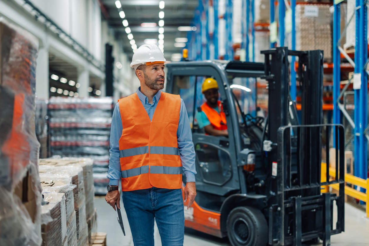 A warehouse manager in an orange safety vest observes a forklift operator navigating through stacks of goods, highlighting the importance of coordination and oversight.