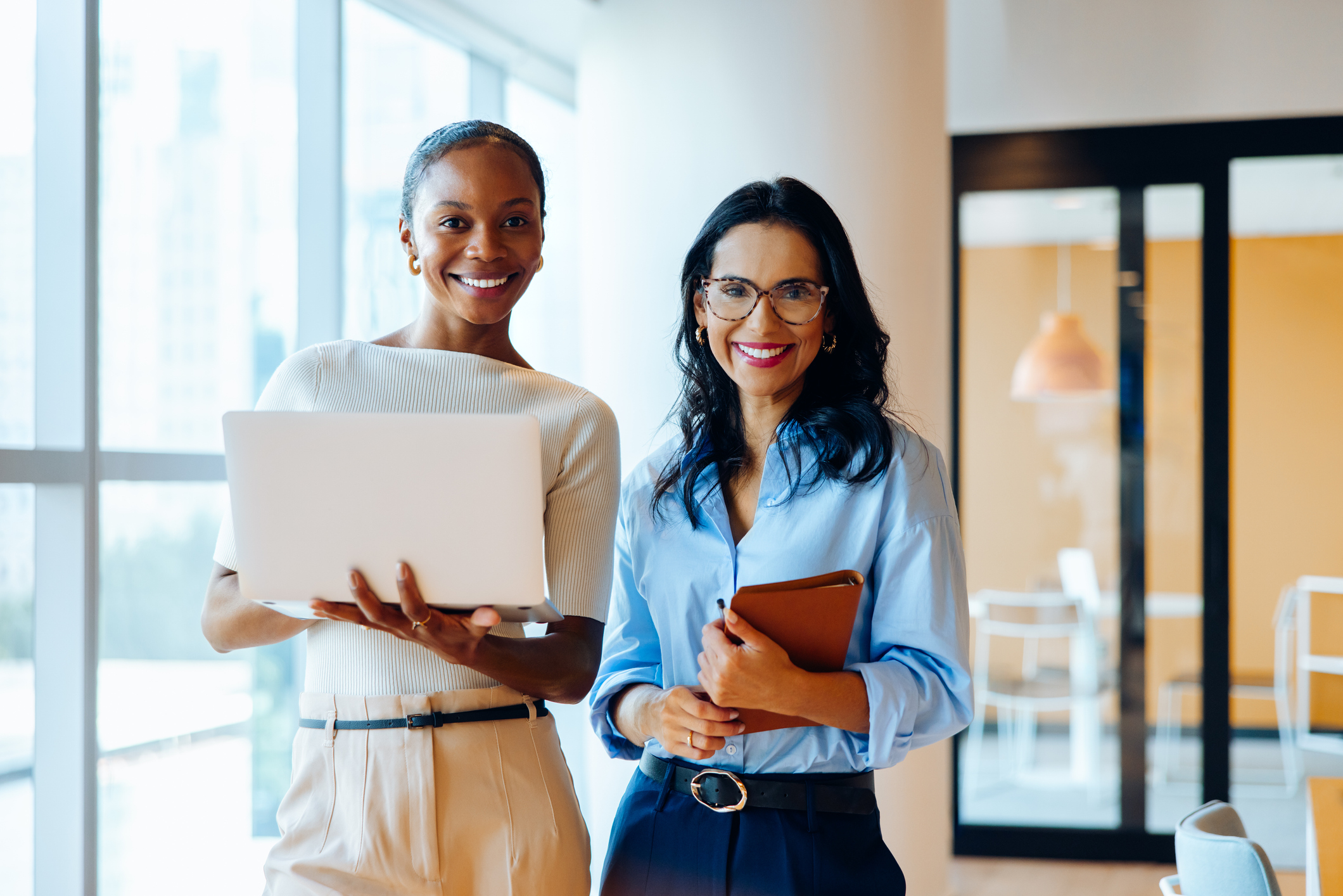 Two confident businesswomen standing in an office space, one holding a laptop, the other holding a notebook. They are smiling and appear professional and friendly.